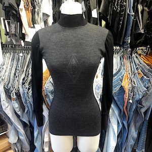 Pode incluir: Uma camisa preta de gola alta e mangas compridas com um logotipo em forma de diamante na frente. A camisa está em um manequim branco.