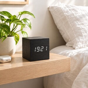 Può includere: Una sveglia digitale nera a forma di cubo con una texture venata del legno. La parte anteriore mostra l'ora 19:21 con numeri LED bianchi luminosi. L'orologio è posizionato su una superficie in legno chiaro accanto a una piccola pianta verde.