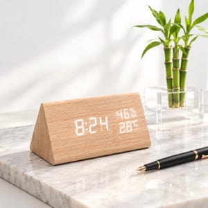 Puede incluir: Un reloj digital triangular de madera clara con una pantalla LED blanca que muestra la hora 8:24, un nivel de humedad del 46% RH y una temperatura de 28 grados Celsius. El reloj tiene un acabado de grano liso y una forma geométrica.