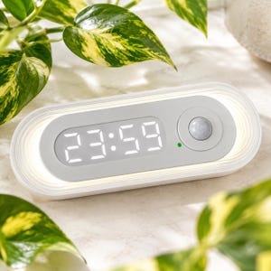 以下が含まれることがあります： 明るい白色のLED数字で23:59と表示された、グレーの中央パネルを持つ楕円形の白いデジタル時計。デバイスの右側には円形のモーションセンサーがあり、小さな緑色のインジケーターライトと照明が統合されています。