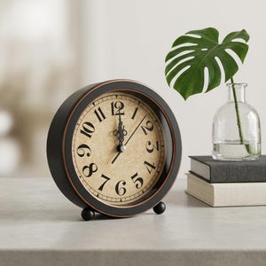 Puede incluir: Un reloj de mesa redondo con marco negro mate y borde interior cobrizo. La esfera presenta un fondo beige similar al pergamino, con grandes números arábigos negros y manecillas de metal negro con un diseño ornamentado.