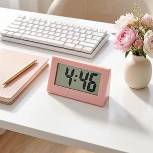 May include: A rectangular digital alarm clock with a matte pink plastic frame and a light gray screen displaying the time 4:46 in bold black digits. The clock has a slanted wedge shape and sits on a flat surface next to a notebook and keyboard.