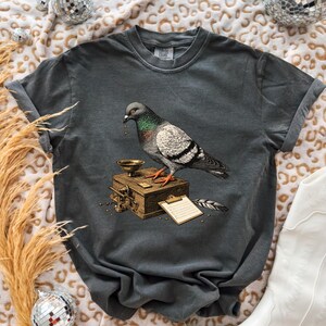 Puede incluir: Una camiseta de manga corta gris carbón con un estampado gráfico de una paloma sobre una caja de madera con un embudo de metal, un pequeño portapapeles y una pluma. La paloma tiene plumas iridiscentes en el cuello.