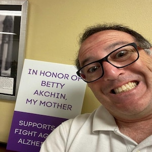 Può includere: Un cartello bianco con testo viola che recita IN HONOR OF BETTY AKCHIN, MY MOTHER e SUPPORTS FIGHT AGAINST ALZHEIMER'S. Il cartello è posizionato accanto a una persona con occhiali dalla montatura nera e una camicia bianca, che sorride.