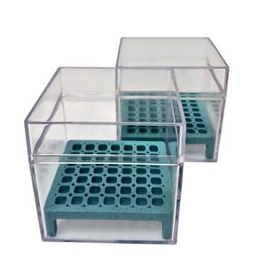 May include: Two clear, square acrylic display boxes, each containing a teal-coloured, rectangular stand with a grid of circular holes. The stands are elevated on short legs and feature a speckled texture, positioned inside the transparent containers.