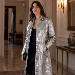 Puede incluir: Un abrigo largo de color plata metalizado con un patrón texturizado de piel de cocodrilo, cuello clásico, mangas largas y cierre frontal de botones. La prenda se lleva sobre una camiseta negra sencilla y pantalones negros, con un corte recto.