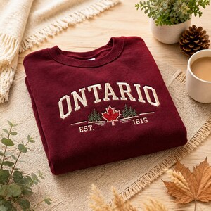 May include: A maroon crewneck sweatshirt featuring embroidered text that reads ONTARIO in a cream-coloured arched font. Below the text is an embroidered red maple leaf flanked by green pine trees and the text EST. 1615 in cream-coloured thread.