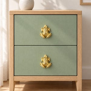 May include: A wooden bedside cabinet with a light oak frame and two sage green drawers. Each drawer features a metallic gold frog-shaped handle centred on the front panel. The piece has four straight legs and a clean, rectangular construction.