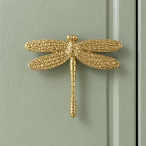 May include: A gold-coloured metal cabinet knob shaped like a dragonfly. The dragonfly features a detailed, textured wing pattern and a segmented body. The knob is mounted horizontally on a flat, sage green surface with a panelled design.