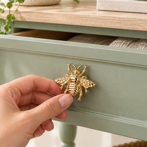 Puede incluir: Un tirador de cajón dorado con forma de abeja que presenta detalles en las alas y los segmentos del cuerpo. Este herraje metálico con forma de insecto tiene un acabado pulido y está diseñado para montarse en muebles para abrir cajones.