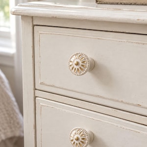 May include: A cream-colored wooden dresser with a distressed finish featuring two round drawer knobs. Each knob has a white, petal-like floral design with a gold-toned center and a beaded edge, mounted on the front of the drawers.