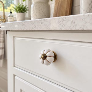 May include: A white ceramic cabinet knob shaped like a pumpkin with a ribbed surface and a metallic gold-toned center accent. The knob is attached to a white drawer front, featuring a rounded, segmented design with a brass-colored base.
