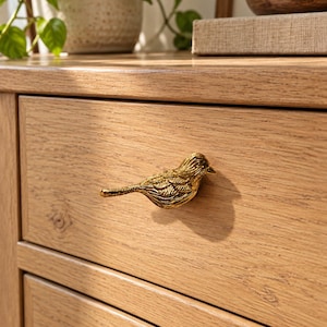 May include: A gold-toned metal bird-shaped drawer pull attached to a light-colored wooden drawer front. The bird figure features detailed engraved lines representing feathers and wings, with a smooth, reflective finish on the surface.