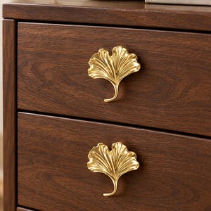 May include: Two gold-colored metal drawer pulls shaped like ginkgo leaves attached to a dark brown wooden cabinet with two drawers. The pulls feature a detailed, textured surface with a metallic finish and a stem-like base pointing downward.