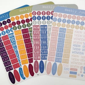 Può includere: Quattro fogli di adesivi per agenda etichettati Weekly Jumbo, con file di cerchi numerati, giorni della settimana in riquadri rettangolari e icone meteo ovali. I fogli includono varie palette di colori come blu, verde, grigio e toni pastello con testo stampato per giorni e attività.