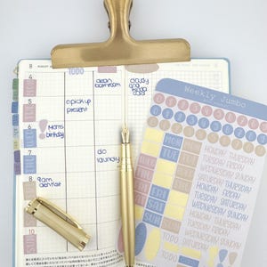 May include: A planner open to an August calendar page with handwritten tasks, held by a brass clip. A gold fountain pen lies on the page next to a sheet of Weekly Jumbo stickers featuring circles, days of the week, and labels in muted pastel tones.