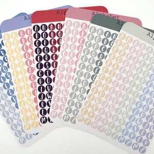 May include: Six rectangular sticker sheets labeled Alphabet, each featuring multiple columns of small circular stickers. Each sticker contains a single white letter of the alphabet in a script font, set against various solid pastel and dark colors.