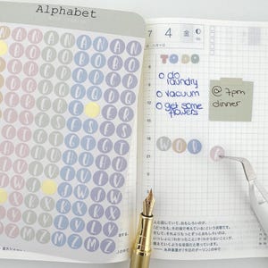 May include: A sheet of circular alphabet stickers in pastel colors featuring letters A through Z, placed on an open planner. The planner page includes handwritten to-do lists, a sticky note with text, a gold fountain pen, and tweezers.