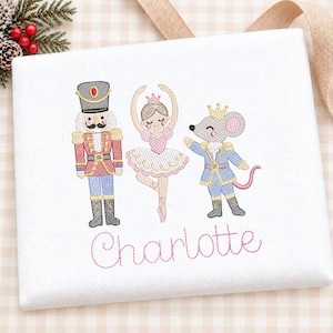 Puede incluir: Un artículo de tela blanca con bordados de un cascanueces con uniforme rojo, una bailarina con vestido rosa y un ratón con corona dorada y chaqueta azul. El nombre Charlotte está bordado debajo en letra cursiva de color rosa.