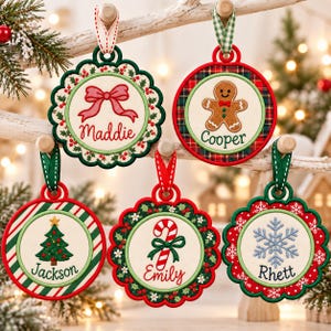 Puede incluir: Cinco adornos navideños redondos para colgar con diseños bordados. Uno presenta un lazo rojo y el nombre Maddie, uno un hombre de jengibre y Cooper, uno un árbol y Jackson, uno un bastón de caramelo y Emily, y uno un copo de nieve y Rhett.