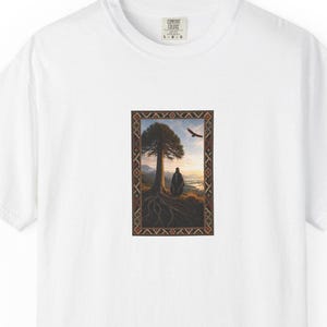 May include: A white short-sleeved t-shirt featuring a rectangular graphic print on the chest. The print depicts a lone figure standing beneath a large tree with visible roots, a bird flying in the sky, and a decorative geometric border frame.