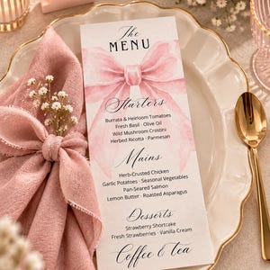 Puede incluir: Una tarjeta de menú rectangular con una ilustración de lazo rosa en acuarela y texto negro que enumera entrantes, platos principales, postres y café y té. El menú está colocado sobre un plato blanco festoneado junto a una servilleta de tela rosa y una cuchara dorada.