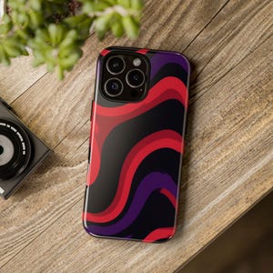 Pode incluir: Uma capa para smartphone com um padrão abstrato e ondulado de linhas fluidas em vermelho vivo, roxo escuro e preto. A capa tem um recorte para um sistema de câmara de lente tripla com as lentes dispostas em formação triangular.