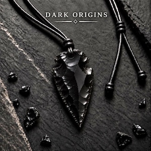 May include: A black obsidian arrowhead pendant suspended on a black cord necklace. The pendant features a sharp, flaked texture and a glossy finish. The text "DARK ORIGINS" is printed in white above the pendant with a small diamond symbol.