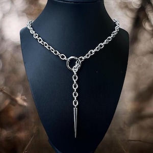 May include: A silver-toned metal chain necklace featuring a circular ring connector at the center. A vertical chain segment hangs from the ring, terminating in a smooth, pointed spike pendant. The necklace is displayed on a black bust form.
