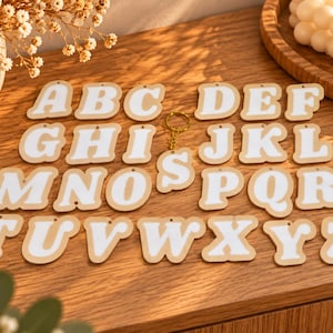 Könnte beinhalten: Eine Sammlung von sechsundzwanzig einzelnen Buchstabenanhängern, die jeweils einen weißen Buchstaben auf hellbraunem Hintergrund zeigen. Die Buchstaben sind in alphabetischer Reihenfolge von A bis Z angeordnet, mit einem goldenen Schlüsselring am Buchstaben S.