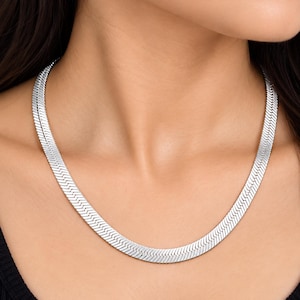 Könnte beinhalten: Eine silberfarbene Fischgräten-Halskette mit flachem, flexiblem Design und einem sich wiederholenden V-förmigen Muster. Die Kette liegt am Hals an und zeigt eine polierte, metallische Oberfläche, die das Licht auf ihren glatten, ineinandergreifenden Gliedern reflektiert.