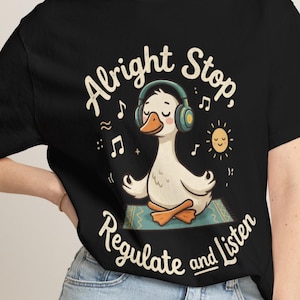 以下が含まれることがあります： 黒いTシャツで、ティール色のヘッドフォンを装着し、模様入りのマットの上で蓮華座のポーズをとる白いアヒルのグラフィックが描かれています。クリーム色のフォントで「Alright Stop, Regulate and Listen」という文字が印刷されており、音符と太陽のイラストが添えられています。