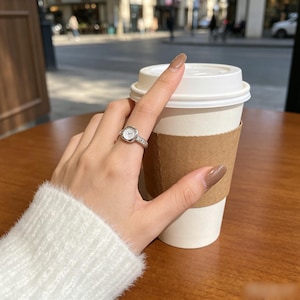 Puede incluir: Un anillo de tono plateado que presenta una pequeña esfera de reloj cuadrada con fondo blanco y manecillas negras. La banda del anillo está adornada con una serie de pequeñas piedras transparentes y brillantes engastadas a lo largo de los lados.