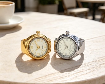 May include: Two ring watches, one in gold-tone and one in silver-tone metal, featuring round faces with white dials, black Roman numeral hour markers, and the word QUARTZ printed on the face. Each watch has a small winding crown at the top.