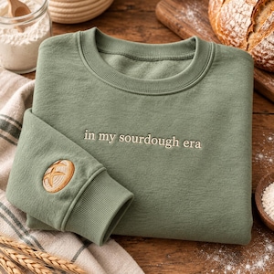May include: A sage green crewneck sweatshirt featuring the embroidered text "in my sourdough era" across the chest and a small, circular embroidered sourdough bread loaf design on the left sleeve cuff.