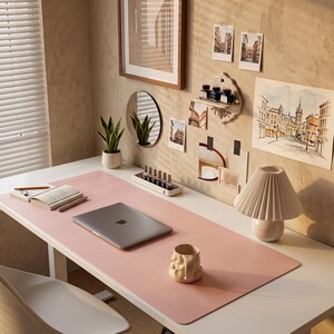 May include: A large rectangular pink desk mat covering a white workspace surface. On the mat sits a closed grey laptop, a beige textured ceramic mug, and a notebook with a pen. A pleated lamp, a small tray with bottles, and a plant are nearby.