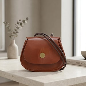 May include: A small, saddle-shaped brown leather crossbody bag with a smooth finish, featuring a rounded flap closure secured by a single circular gold-toned metal button and a thin, dark brown leather shoulder strap.