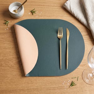 Pode incluir: Um lugar americano verde-escuro com uma borda arredondada e um canto dobrado revelando um lado inferior em tom pêssego claro. Um garfo e uma faca de metal dourado estão colocados sobre a superfície. O item possui uma textura semelhante ao couro.
