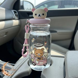 Puede incluir: Una botella de agua de vidrio transparente con una tapa rosa claro rematada por una cabeza de oso beige. La botella cuenta con una correa de transporte de silicona rosa y un estampado gráfico de personajes Rilakkuma en un scooter con el texto Always with Rilakkuma.