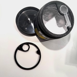 Könnte beinhalten: Ein schwarzer Kunststoffbehälter mit einem gelben Etikett mit der Aufschrift ESN, einem durchsichtigen Deckel, einem separaten schwarzen Ringeinsatz und einem kleinen transparenten Kunststofflöffel in dem mit weißem Pulver gefüllten Behälter.