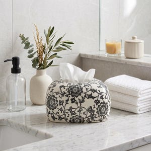 May include: A rectangular ceramic tissue box cover featuring a black floral pattern on a cream background. The box has rounded edges and holds a stack of white tissues. The design displays intricate botanical motifs with swirling vines and flowers.