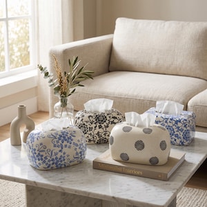 May include: Four oval-shaped ceramic tissue box covers with various patterns including blue and white floral, black and white floral, and cream with black polka dots. The covers are arranged on a marble table alongside a book titled Minimalist Interiors.