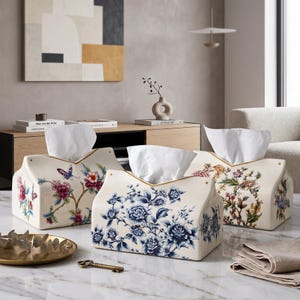 Könnte beinhalten: Drei cremefarbene Taschentuchboxen aus Keramik in Hausform. Eine zeigt blaue Blumenmuster, eine weitere bunte Blumen mit einem Schmetterling und die dritte aufwendige botanische Designs, jeweils bestückt mit weißen Papiertüchern.