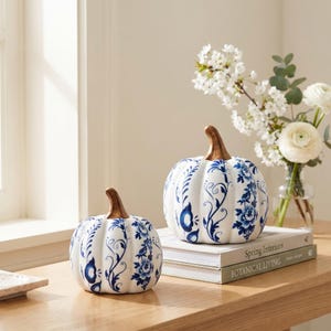 May include: Two white ceramic pumpkins featuring blue floral patterns and metallic bronze stems. The smaller pumpkin sits on the surface, while the larger pumpkin rests on top of two stacked books titled Spring Interiors and Botanical Living.