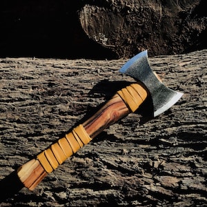 May include: A double-bit axe featuring a dark, textured metal head with polished edges. The handle is crafted from polished wood with prominent grain patterns, wrapped in tan leather strips at the base and near the axe head.