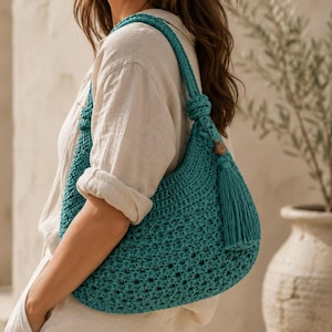 May include: A teal crochet shoulder bag featuring an intricate openwork pattern, a single knotted strap, and a decorative tassel accent attached with a small wooden bead. The bag has a crescent shape and is crafted from a textured yarn material.