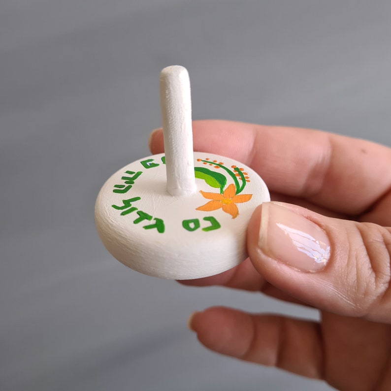 hand holding white wooden dreidel with orange flower green leaves and Nes Gadol Haya Po writing in green