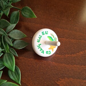 white wooden dreidel with orange flower green leaves and Nes Gadol Haya Po writing in green