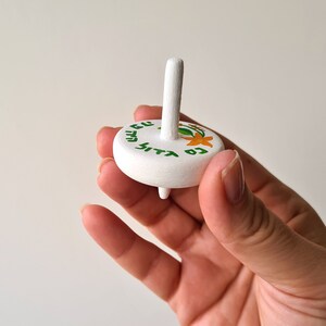 hand holding a white wooden dreidel with orange flower green leaves and Nes Gadol Haya Po writing in green