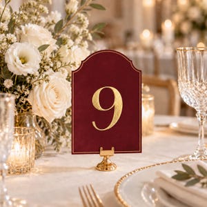 Peut inclure: Une carte de numéro de table bordeaux en forme d'arche avec un grand chiffre 9 en feuille d'or métallique au centre. La carte est maintenue droite par un petit support circulaire en métal doré, avec une fine bordure dorée.